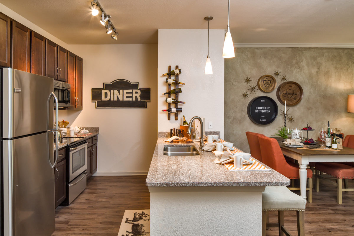 Model apartment kitchen and dining area with stainless steel appliances and fixtures, overhead lighting, granite countertop, and hardwood flooring