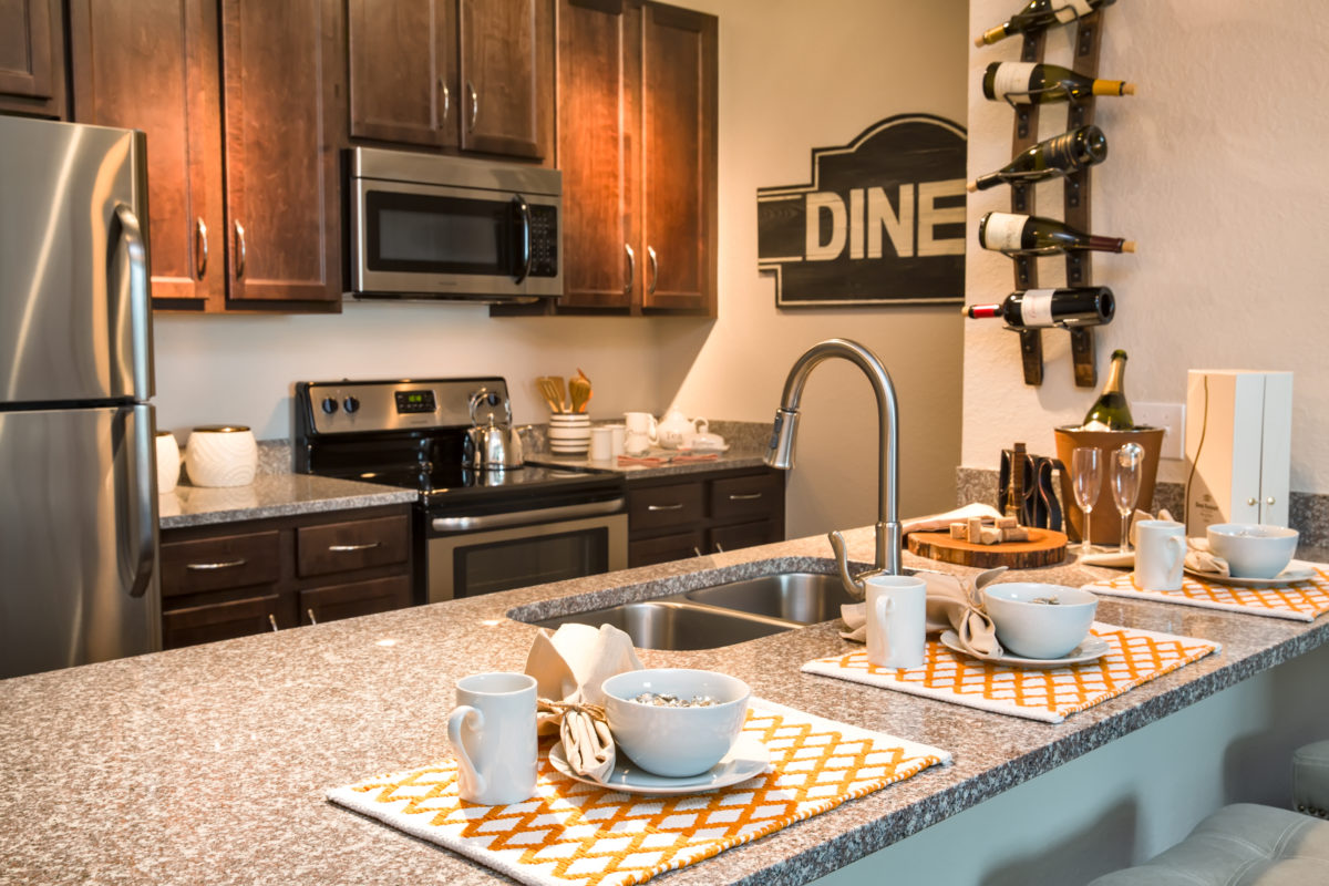 View of model apartment kitchen with granite countertop, bar area, double sink faucet, and stainless steel fridge, microwave, and oven with glass stove