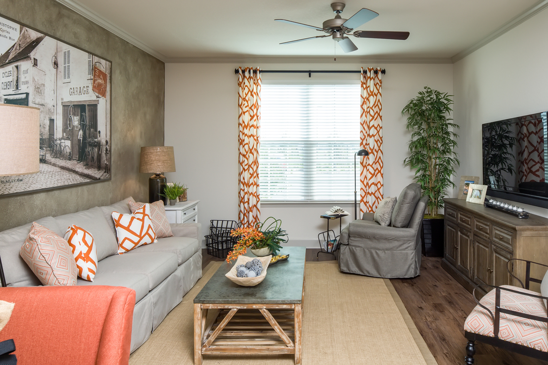 Model apartment living room with ceiling fan, accent wall, hardwood flooring, and window with wood blinds. Example furniture includes couch, armchair, accent chair, TV, and coffee table