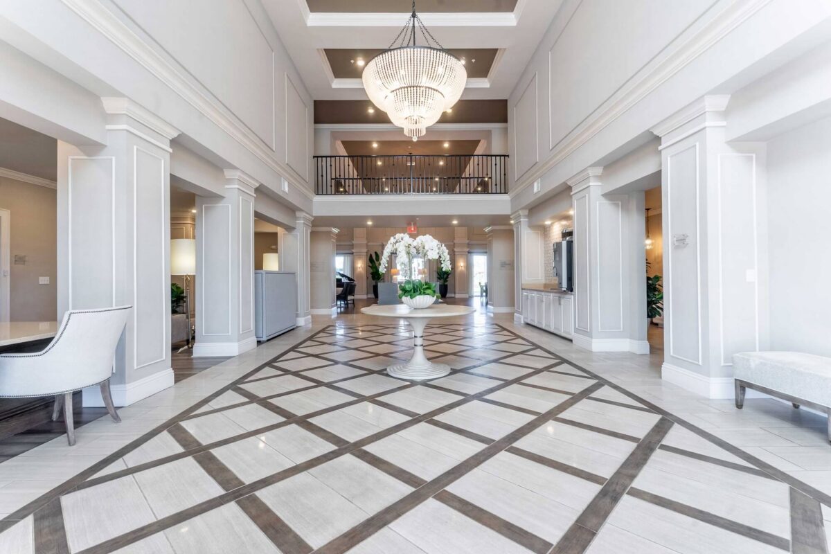 Bright, elegant hotel lobby with tall ceilings, a large chandelier, white columns, a central round table with flowers, patterned tile flooring, and a second-floor balcony overlooking the space.