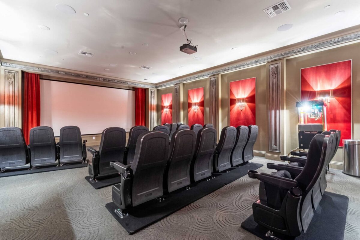 A luxurious home theater room with three rows of black reclining seats facing a large projector screen, red curtains and red wall panels, and a projector mounted on the ceiling.