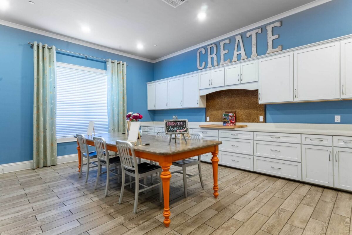 A creative workspace with blue walls, white cabinets, a large wooden table with chairs, a window with curtains, and a sign reading CREATE above the cabinets. Art supplies and flowers are on the table.