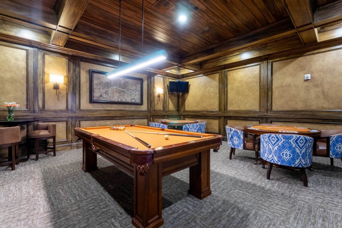 A cozy game room with a wooden pool table, orange felt top, two blue patterned armchairs around a card table, wood-paneled walls, a wall-mounted TV, and a framed map on the back wall.