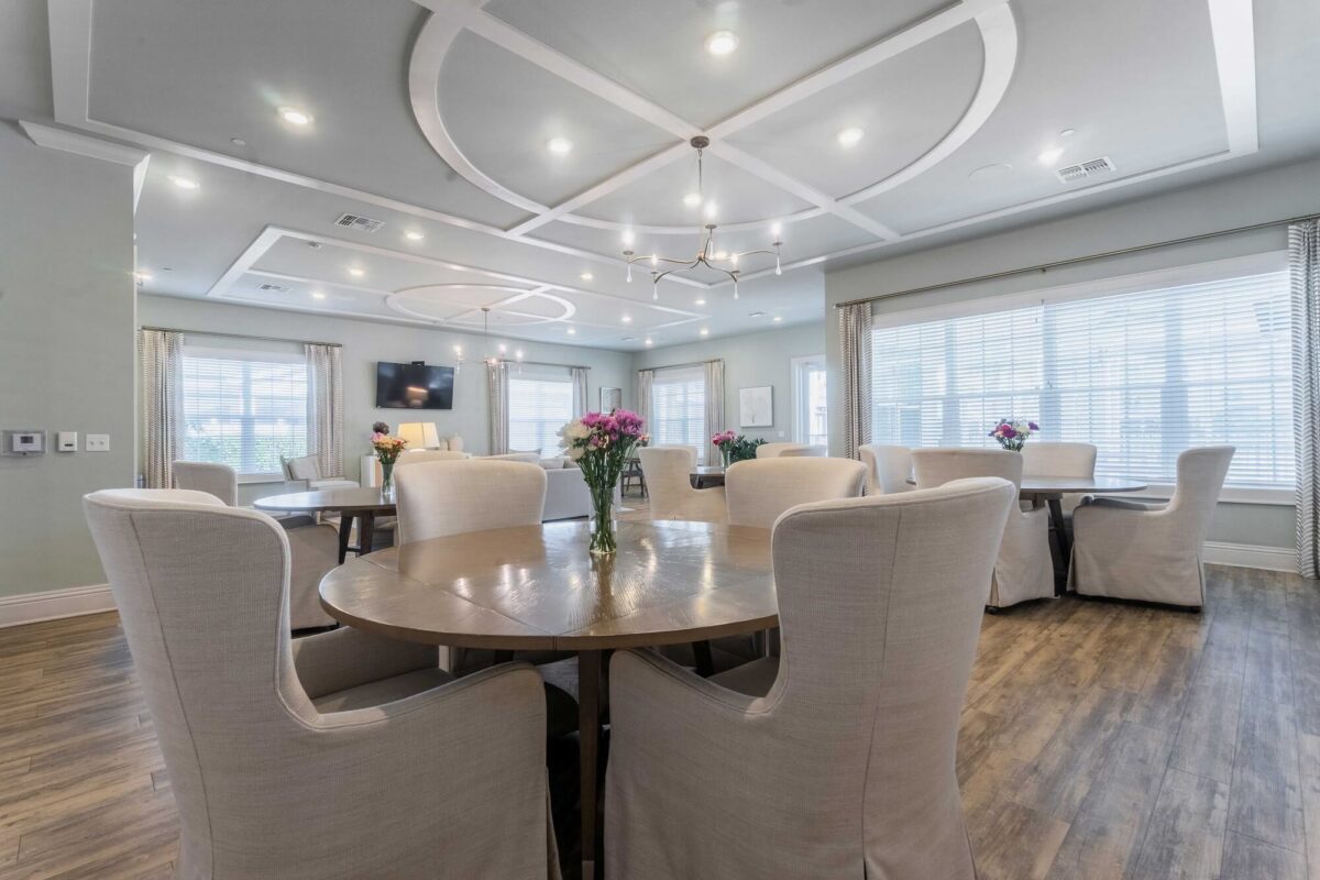 A spacious, modern dining room with round tables, cream-colored upholstered chairs, wood flooring, large windows with blinds, and decorative ceiling lights. Vases with flowers sit on each table.