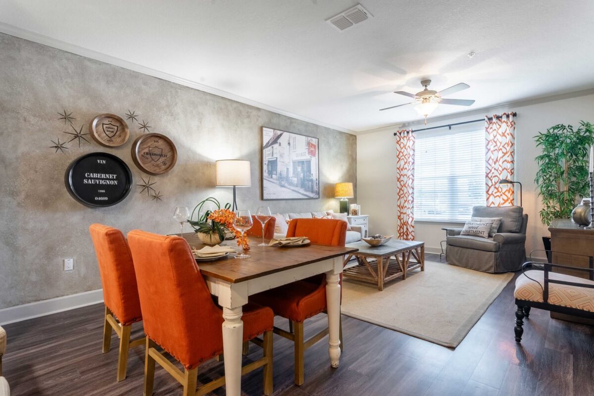 A modern living and dining area features a wooden table with four orange chairs, a cozy seating area with a gray armchair and sofa, large window with patterned curtains, wall art, and decorative accents.
