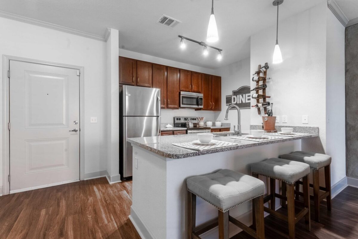 Modern kitchen with stainless steel appliances, wooden cabinets, granite countertops, three cushioned bar stools, pendant lighting, and a decorative DINE sign on the wall above the sink.