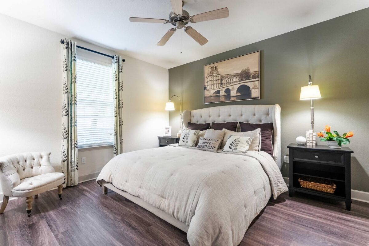 A cozy bedroom with a large beige bed, decorative pillows, two black nightstands with lamps, a gray accent wall, a window with curtains, a white tufted chair, and a ceiling fan. A framed artwork hangs above the bed.