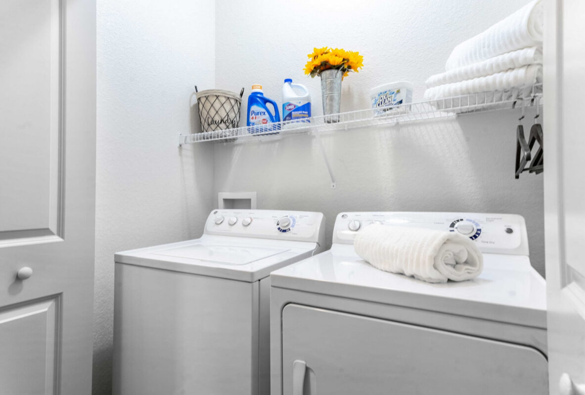 A bright laundry room with a white washer and dryer, folded white towels, laundry supplies, a basket, and a vase of yellow flowers on a wire shelf above the machines.