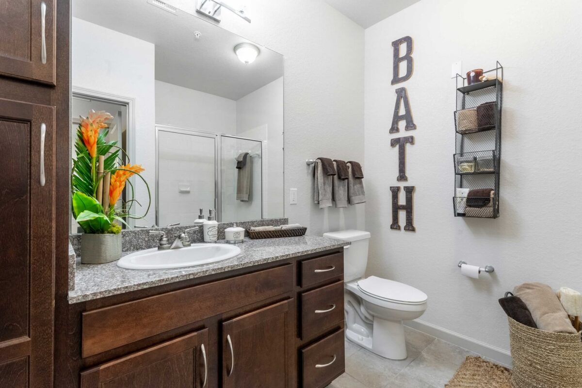 Modern bathroom with a granite countertop, sink, dark wood cabinets, and a toilet. Towels are neatly arranged, and “BATH” is spelled out on the wall. A basket, shelves, and a vase with orange flowers add decorative touches.