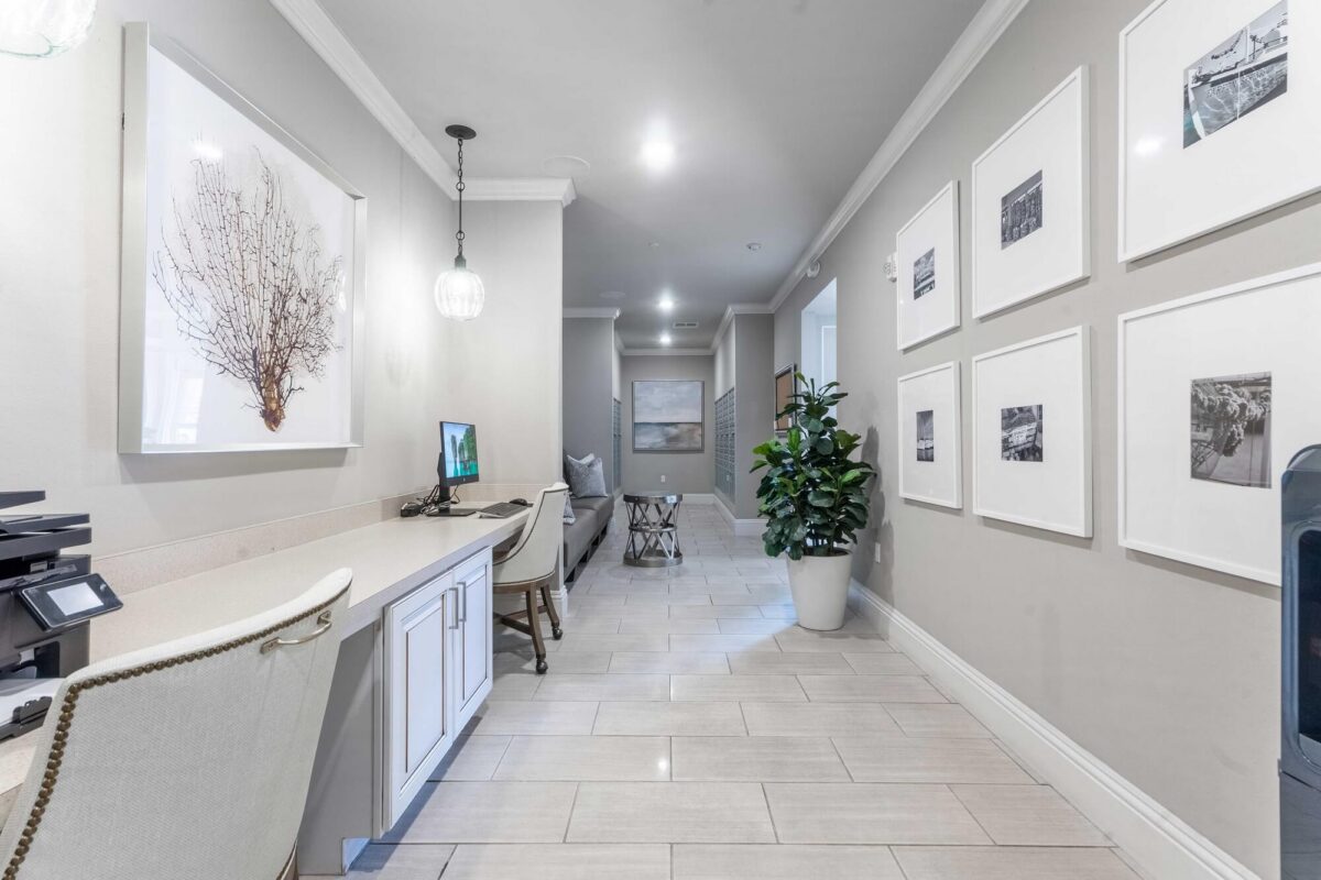 A modern office hallway with light tile flooring, two white desks with chairs and computers on the left, framed art on the walls, and a large potted plant near the hallway’s end. The space is bright and well-lit.