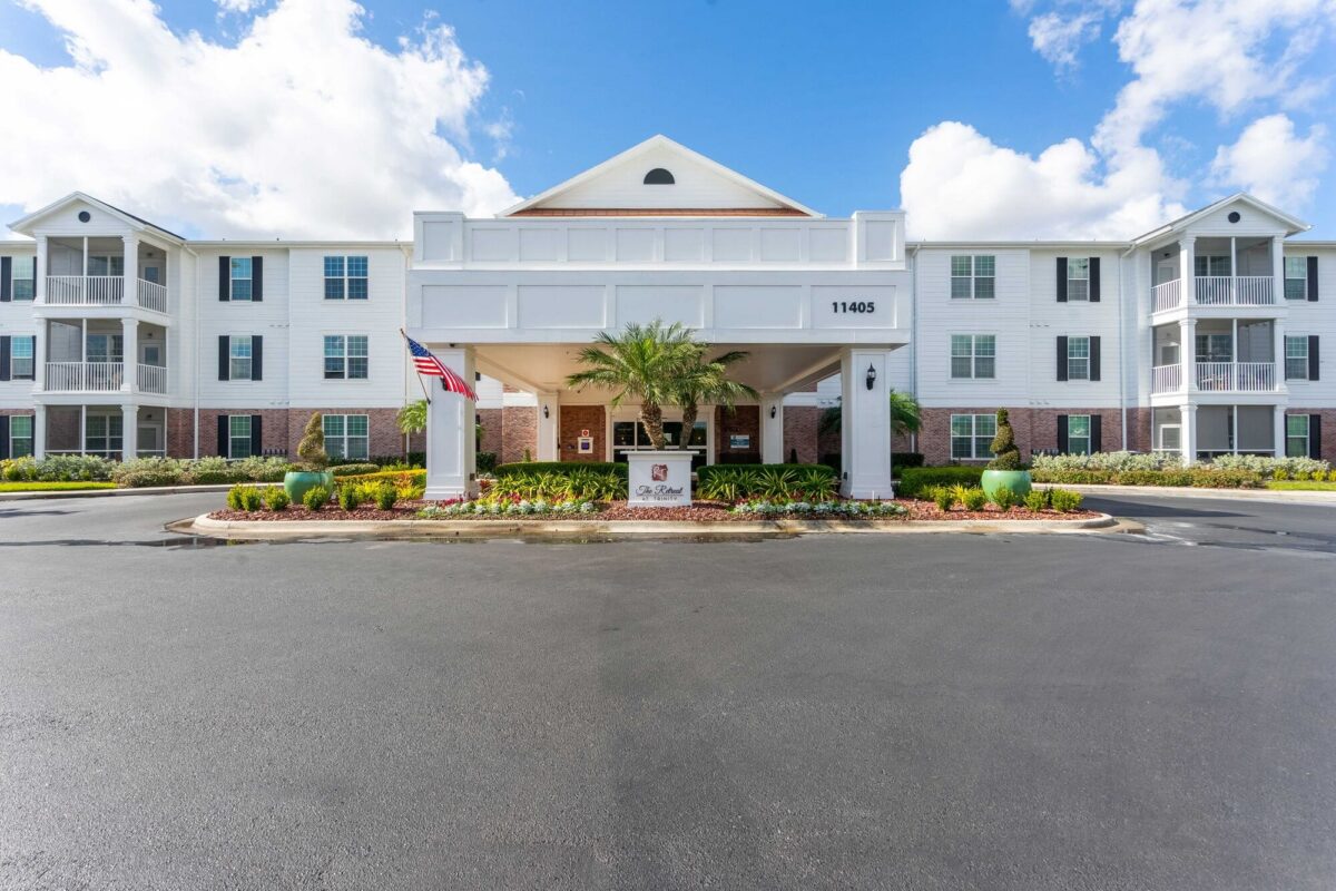 A large, three-story white building with a covered entrance, American flag, palm trees, landscaping, and a sign reading The Colonnade in front; bright blue sky with clouds overhead.