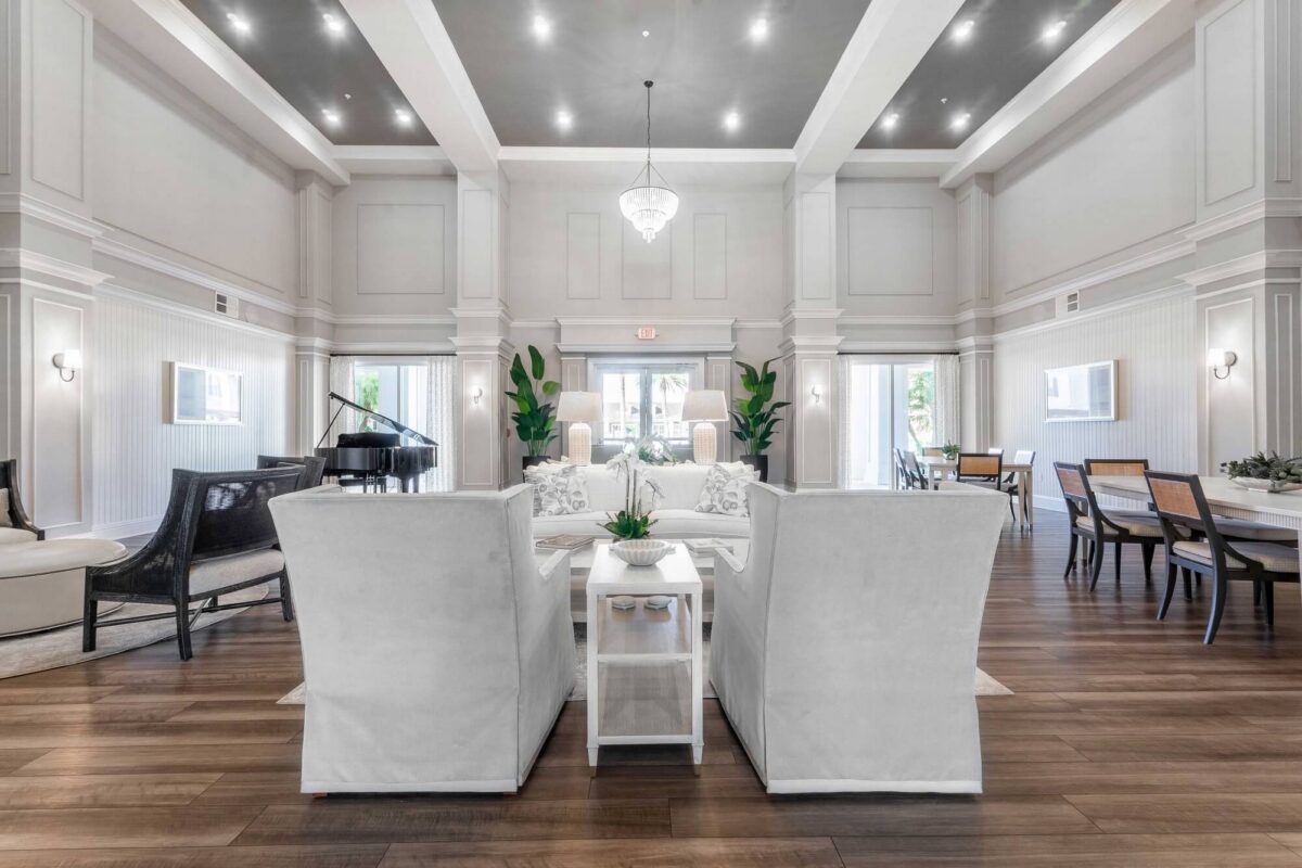 A spacious, elegant living room with high ceilings, white walls, modern furniture, a chandelier, wood flooring, large windows, a grand piano, and green plants for decoration.