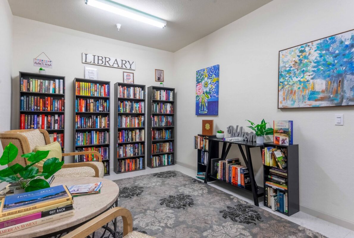 Cozy library room with three tall bookshelves filled with colorful books, a small bookshelf, two armchairs near a round table with books, and decorative paintings on cream walls. A rug covers part of the floor.