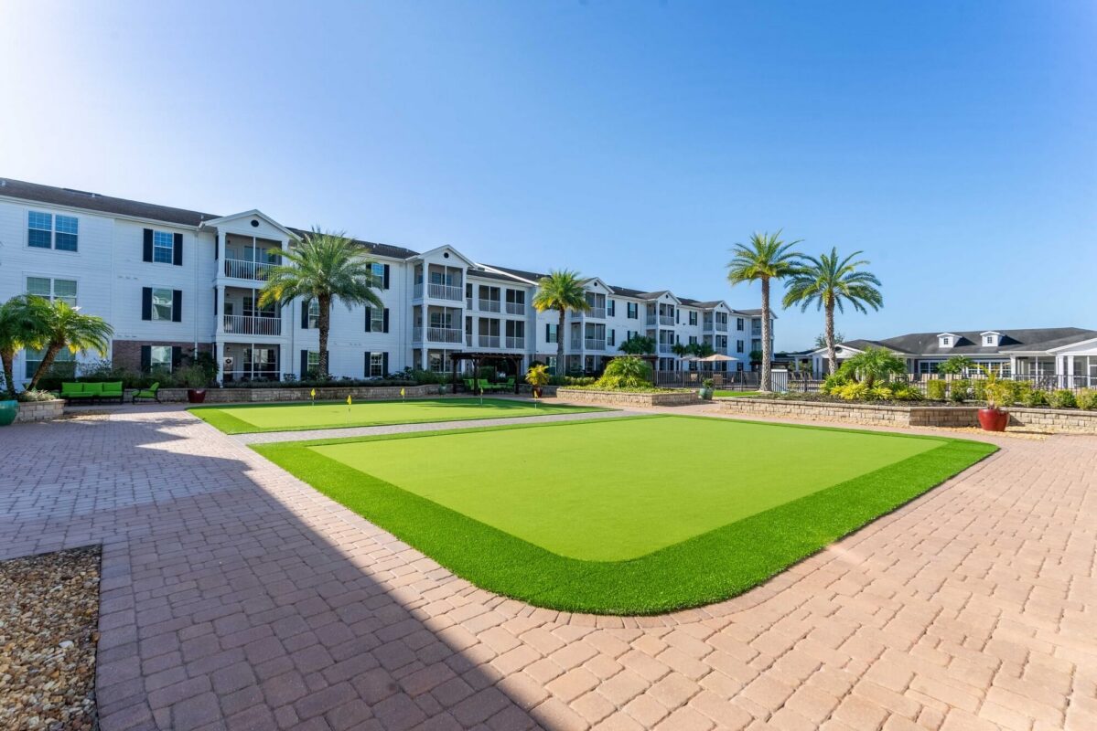 A modern apartment complex with white buildings, balconies, and black roofs overlooks a green lawn area with palm trees and paving stones under a clear blue sky.