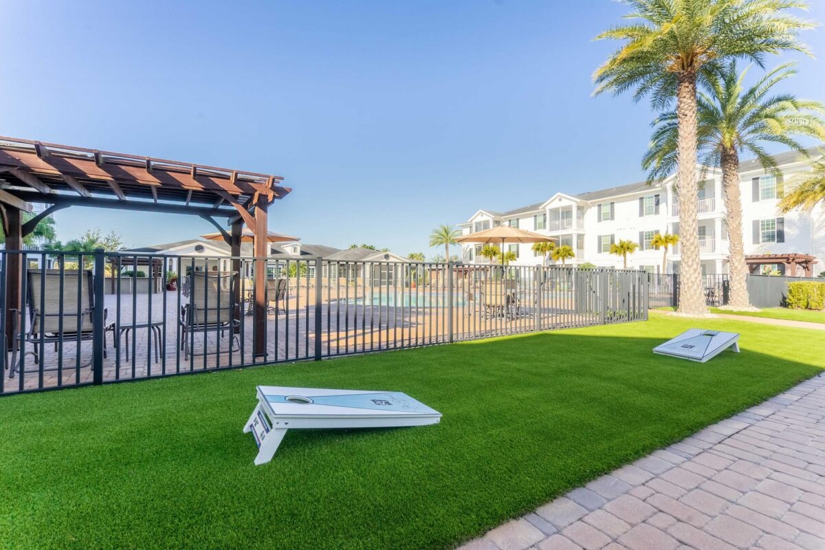 A well-maintained outdoor area with artificial grass, two cornhole boards, a fenced swimming pool, palm trees, a pergola with seating, and a white multi-story building in the background under a clear blue sky.