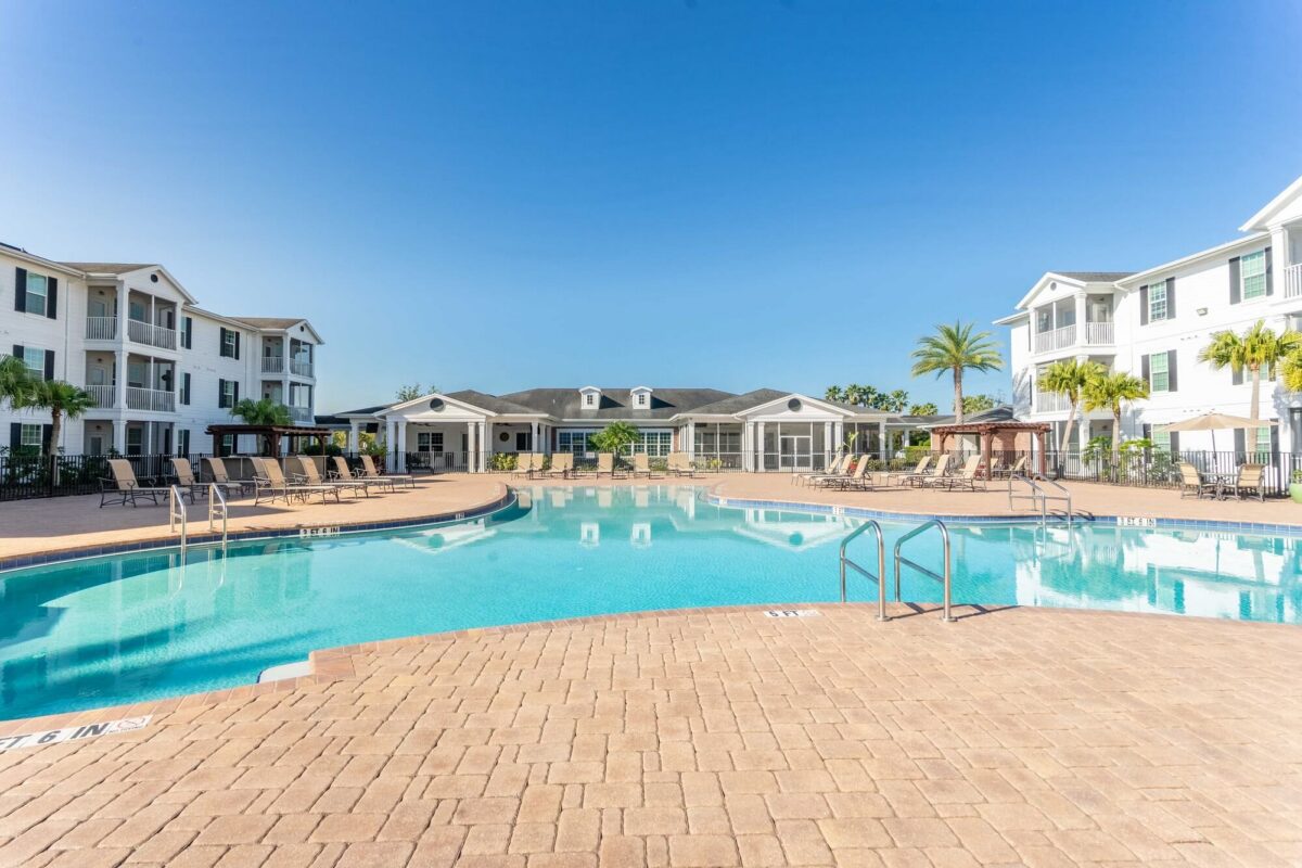 Large outdoor swimming pool surrounded by lounge chairs, umbrellas, and three-story white apartment buildings with balconies under a clear blue sky.