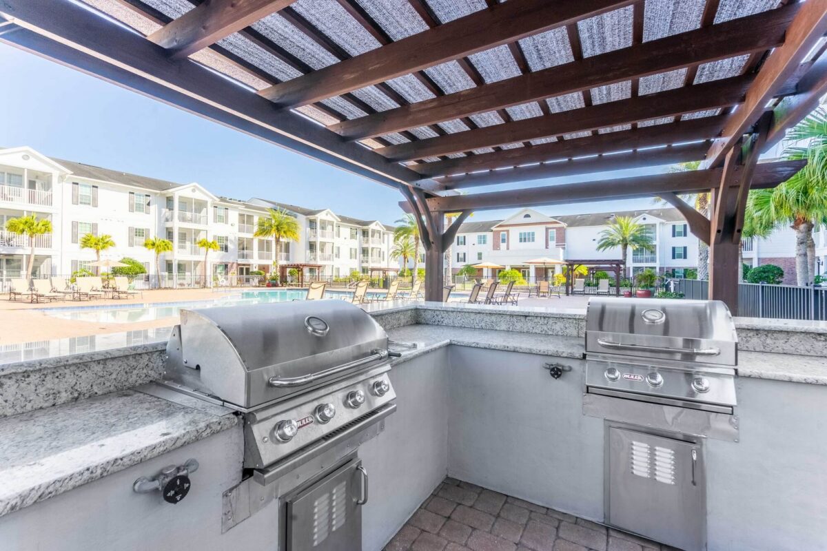 Outdoor grill station with two stainless steel barbeque grills under a wooden pergola, overlooking a sunny pool area with lounge chairs, palm trees, and modern apartment buildings in the background.