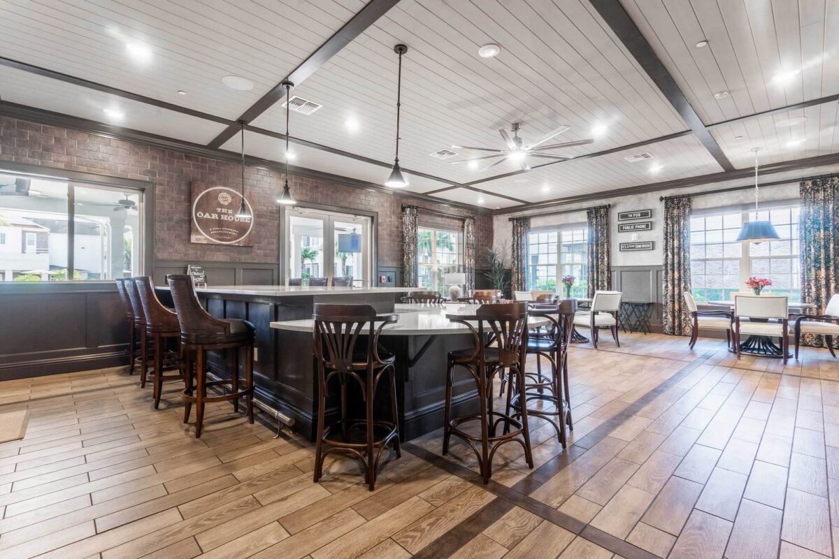 Spacious, well-lit communal room with a large central island, high-backed chairs, tables, and windows letting in natural light. The room features wood floors, exposed beams, and decorative accents.